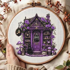Könnte beinhalten: Ein runder Holzstickrahmen mit einem Kreuzstichmuster eines violetten Ladens mit einer schwarzen Katze vor der Tür. Der Laden zeigt Regale voller Flaschen, violetter Rosen, Pilze und ein Schild mit der Aufschrift OPEN.
