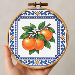 May include: A cross-stitch embroidery hoop depicting a branch of oranges with green leaves and white flowers. The design is framed by a blue and white decorative border. The wooden hoop adds a rustic touch. The design is set against a white background.