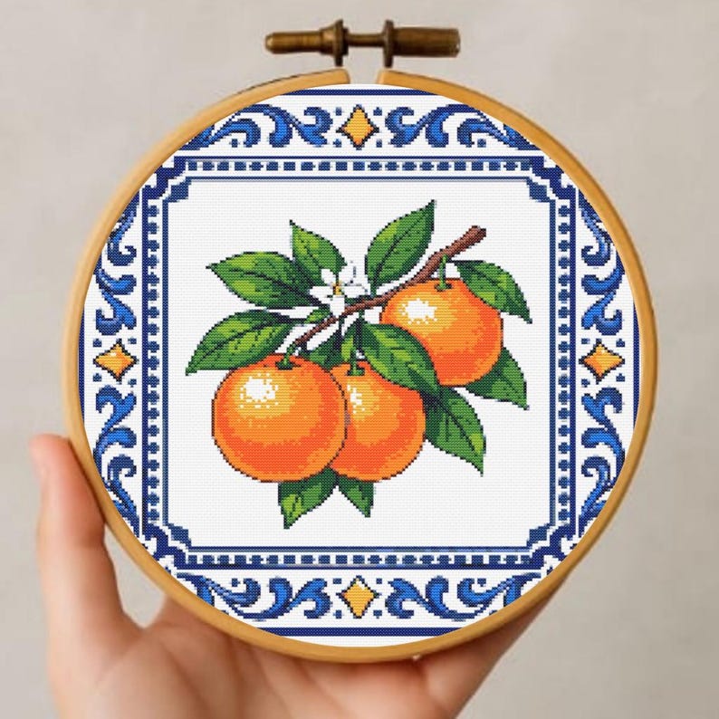 May include: A cross-stitch embroidery hoop depicting a branch of oranges with green leaves and white flowers. The design is framed by a blue and white decorative border. The wooden hoop adds a rustic touch. The design is set against a white background.