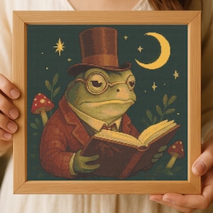 May include: A framed cross-stitch artwork depicting a frog in a top hat and spectacles, reading a book. The frog is wearing a suit and is surrounded by mushrooms, stars, and a crescent moon. The colour palette includes shades of green, brown, and red.