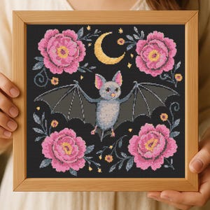 Könnte beinhalten: Kreuzstich-Kunstwerk in einem Holzrahmen. Es zeigt eine graue Fledermaus mit ausgebreiteten Flügeln, einen Halbmond und rosa Rosen auf schwarzem Hintergrund. Das Design hat ein skurriles, gotisches Thema mit floralen und himmlischen Elementen.