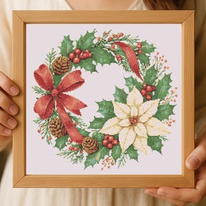 May include: Cross-stitch Christmas wreath in a wooden frame. The wreath features green holly leaves, red berries, pinecones, and a large red bow. A cream-colored poinsettia is also included in the design. The artwork is held by a person.