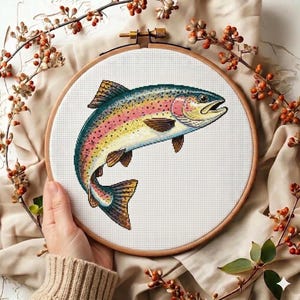 May include: A colorful cross-stitch of a rainbow trout displayed in a wooden embroidery hoop. The fish features shades of blue, pink, yellow, and brown. The hoop is held by a person's hand, with a beige sweater sleeve visible. Decorative branches surround the hoop.