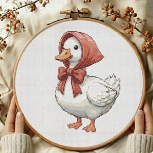 Könnte beinhalten: Kreuzstich-Stickerei einer weißen Ente mit roter Haube und Schleife. Die Ente hat einen orangefarbenen Schnabel und Füße. Die Stickerei befindet sich in einem Holzreifen und zeigt ein charmantes, handgefertigtes Design.