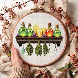 May include: Cross-stitch embroidery in a wooden hoop. The design features a shelf with potion bottles, candles, and crystals. Below the shelf are hanging herbs and crystals. The color palette includes green, orange, yellow, and purple.