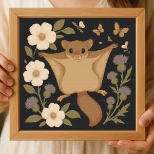 May include: A framed cross-stitch featuring a cartoon sugar glider with outstretched arms, surrounded by cream-colored flowers, purple thistles, butterflies, and green leaves on a black background. The frame is made of light brown wood.