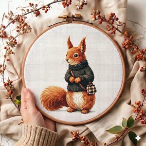May include: A cross-stitch embroidery hoop featuring a detailed image of a squirrel wearing a dark sweater and carrying a plaid bag. The squirrel has orange fur and is set against a white background. The wooden hoop is held by a hand.