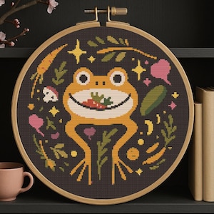 May include: A cross-stitch embroidery hoop featuring a smiling orange frog holding food, surrounded by plants and stars on a dark background. The wooden hoop is round. A pink mug and books are in the background.