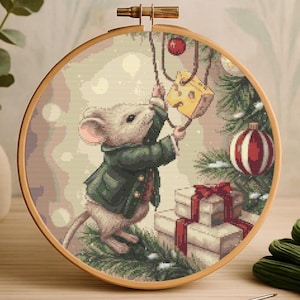 May include: Cross-stitch embroidery in a wooden hoop features a mouse in a green coat decorating a Christmas tree with a cheese ornament. Presents with red ribbons are at the base of the tree. The background is a soft, neutral color.