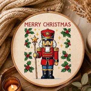Könnte beinhalten: Ein runder Holzstickrahmen mit einem Kreuzstichmotiv eines Nussknackers in roter Uniform, der einen goldenen Sternenstab hält. Der Text MERRY CHRISTMAS ist in Rot darüber gestickt, umgeben von grünen Stechpalmenblättern und roten Beeren.