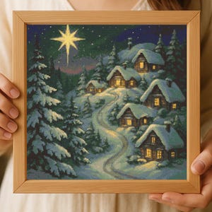 May include: A framed cross-stitch depicting a snowy village scene at night. The artwork features illuminated houses nestled in a snow-covered landscape, a winding road, and a bright star in the sky. The frame is made of light-colored wood.