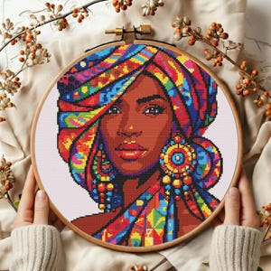 May include: A cross-stitch portrait of a woman with dark skin wearing a colorful head wrap and large earrings. The embroidery is set in a wooden hoop, showcasing a vibrant design with shades of red, blue, yellow, and green.
