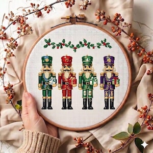 Pode incluir: Um bastidor de bordado em ponto cruz com quatro soldados quebra-nozes coloridos. Cada soldado está vestido com uma cor diferente: verde, vermelho, verde e roxo. Eles estão segurando presentes e são decorados com detalhes dourados. Uma guirlanda de azevinho e bagas está acima deles.