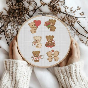 May include: A cross-stitch embroidery hoop featuring several teddy bears in various poses. Some bears hold a heart-shaped balloon, flowers, and a sign that says "LOVE". The bears are in shades of brown and beige, with red accents.