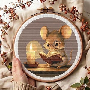 May include: A circular embroidery hoop containing a cross-stitch pattern of a light brown mouse wearing round glasses, reading an open book beside a lit candle. The mouse is positioned on a wooden surface against a solid brown background.