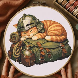 May include: Cross-stitch embroidery of a sleeping ginger tabby cat wearing a medieval helmet, resting on a green backpack with a rolled-up blanket and canteen. The embroidery is set in a wooden hoop.