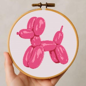 Pode incluir: Um bastidor de bordado em ponto cruz rosa com um desenho de cão balão. O cão é feito de fio rosa e está sobre um fundo branco. O bastidor de madeira é segurado por uma mão.