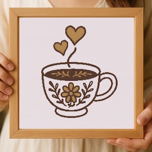 May include: Cross-stitch art in a wooden frame featuring a brown coffee cup with a floral design and two hearts rising above. The cup is filled with coffee and has a handle. The background is white.