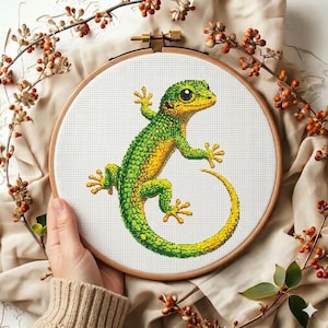 Peut inclure: Broderie au point de croix d'un gecko vert et jaune dans un cercle en bois. Le gecko est détaillé avec des nuances de vert et de jaune. Le fond est blanc et le cercle est tenu par une personne. Des branches décoratives avec des baies oranges entourent le cercle.