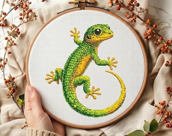 May include: A cross-stitch embroidery of a green and yellow gecko inside a wooden hoop. The gecko is detailed with various shades of green and yellow. The background is white, and the hoop is held by a person. Decorative branches with orange berries surround the hoop.