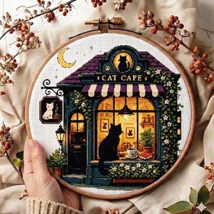 May include: A circular embroidery hoop containing a cross-stitch pattern of a cat cafe building. The design features a black cat in the window, a purple roof, a striped awning, and a sign reading CAT CAFE. The scene includes a crescent moon.