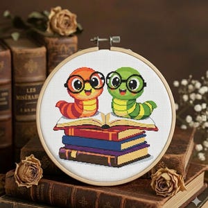 Könnte beinhalten: Kreuzstich-Stickerei in einem Holzreifen mit zwei Comic-Büchern, die eine Brille tragen und auf einem Stapel Bücher sitzen. Ein Wurm ist orange und rot, der andere grün und gelb. Die Bücher sind in verschiedenen Farben.