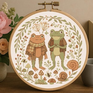 Może przedstawiać: Obręcz do haftu krzyżykowego z dwoma antropomorficznymi żabami w jesiennych strojach. Wzór obejmuje wieniec z liści, grzybów i ślimaków. Paleta kolorów obejmuje odcienie brązu, zieleni i pomarańczy.