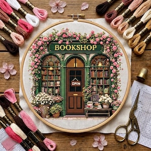 May include: A cross-stitch embroidery in a wooden hoop depicting a green bookshop storefront with the text BOOKSHOP and OPEN, surrounded by pink flowers and greenery. The piece is displayed with various coloured embroidery floss, scissors, and a thimble.