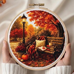 Puede incluir: Un bastidor de bordado circular con un patrón de punto de cruz de una escena de parque en otoño. El diseño muestra un banco de madera con un cojín, libros y una taza, bajo un árbol de hojas naranjas, junto a una farola negra y calabazas.