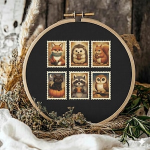 May include: A black embroidery hoop with six postage stamp-style images of woodland creatures. The images include a fox, hedgehog, squirrel, cat, raccoon, and owl. The embroidery hoop is set against a rustic wooden background with dried plants.