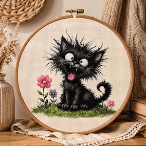Puede incluir: Un bordado de punto de cruz en un bastidor de madera redondo que muestra un gato negro esponjoso con ojos grandes y la boca abierta sentado sobre hierba verde. El diseño incluye una flor rosa, una flor morada pequeña y una flor rosa diminuta.
