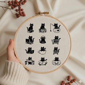 May include: A round embroidery hoop featuring a cross-stitch design of black cats in various poses. The cats are depicted sitting on books, wearing headphones, and in a shopping cart. The design is set against a cream-colored fabric.