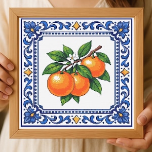 May include: Cross-stitch artwork of three oranges on a branch with green leaves and white blossoms. The oranges are framed by a blue and white decorative border with floral and geometric patterns. The artwork is in a wooden frame.