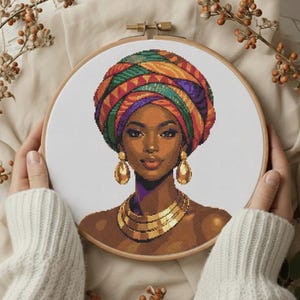 May include: A cross-stitch portrait of a woman wearing a colorful head wrap, gold earrings, and a gold necklace. The embroidery is set in a wooden hoop. The woman has brown skin and is smiling.