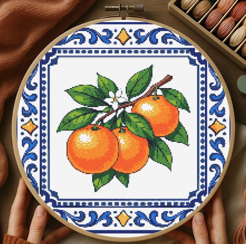 May include: Cross-stitch embroidery of oranges on a branch with green leaves and white flowers. The design is framed by a blue and white border within a wooden hoop. The embroidery features a detailed, pixelated style.