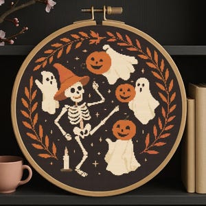 Peut inclure: Une broderie au point de croix sur le thème d'Halloween, dans un cercle en bois. Le motif représente un squelette portant un chapeau de sorcière, entouré de fantômes et de citrouilles, sur fond sombre avec une bordure de feuilles.