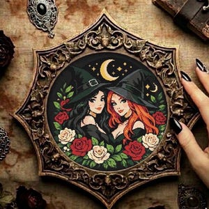 May include: A framed cross-stitch featuring two witches with black and red hair, wearing black hats, surrounded by red and cream roses. The background is black with a crescent moon and stars. The frame is ornate, with a dark, antique finish.
