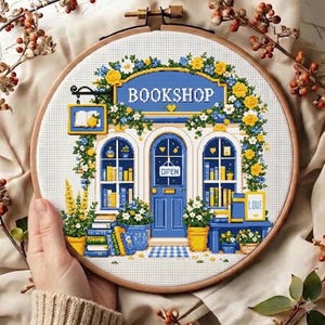 May include: A circular wooden embroidery hoop containing a cross-stitch pattern of a bookshop storefront. The design features a blue door with an OPEN sign, large windows displaying books, and yellow and blue floral accents. The text BOOKSHOP is visible.
