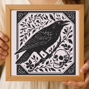 May include: A framed cross-stitch featuring a black raven perched on a branch, with a skull and foliage. The artwork is set against a white background and framed in a light brown wooden frame. The design is a gothic style.