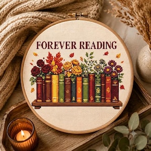 May include: A circular embroidery hoop containing a cross-stitch pattern on cream fabric. The design features a row of colourful books on a shelf with autumn flowers and leaves above them. The text FOREVER READING is stitched in dark red letters.