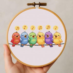 Può includere: Un telaio da ricamo a punto croce con sei uccelli colorati su un filo, ognuno con note musicali sopra la testa. Gli uccelli sono rossi, blu, verdi, gialli, viola e gialli. Il ricamo è in un telaio di legno.