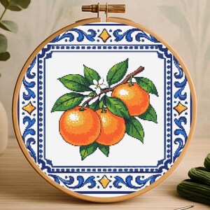 May include: Cross-stitch embroidery of oranges on a branch with green leaves and white blossoms. The design is framed in a wooden hoop with a blue and white border. The oranges are a vibrant orange colour.