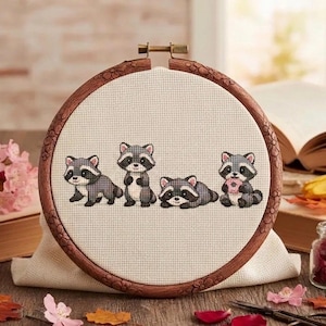 May include: A circular wooden embroidery hoop containing a piece of cream-colored Aida fabric. The fabric features a cross-stitch pattern of four small raccoons in various poses, including one holding a pink donut, rendered in gray and black thread.