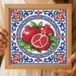 May include: A framed cross-stitch artwork featuring three pomegranates. Two whole pomegranates and one cut open, revealing the seeds, are surrounded by green leaves. The design is set within a circular pattern with blue and red floral motifs.