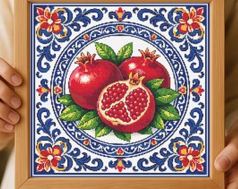 May include: A framed cross-stitch artwork depicting three pomegranates. Two whole pomegranates and one cut open, showing seeds, are surrounded by green leaves. The design is set within a circular pattern with blue and red floral motifs.