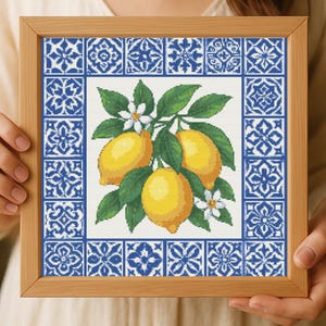 Peut inclure: Broderie au point de croix de citrons sur une branche avec des feuilles vertes et des fleurs blanches. Les citrons sont jaunes et les feuilles sont vertes. L'œuvre est encadrée dans un cadre en bois avec une bordure de carreaux bleus et blancs.