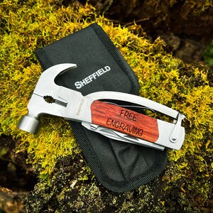 May include: A multi-tool with a hammer head, pliers, and a knife blade. The tool has a wooden handle with the words "FREE ENGRAVING" engraved on it. The tool is sitting on a bed of green moss.