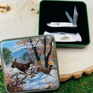Puede incluir: Un set Remington Whitetails Limited Edition que incluye dos navajas plegables con mangos color crema y grabados de ciervos, presentadas en una lata de metal verde con una tapa que muestra un bosque con dos ciervos y el texto Remington.