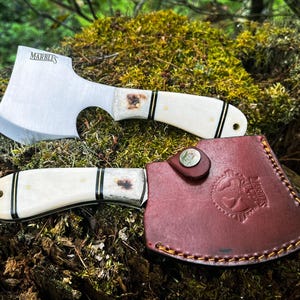 May include: A small, white-handled axe with a brown leather sheath. The axe has a black and gold inlay on the handle and the text "MARBLES" on the blade.