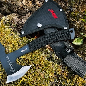 May include: Black tactical tomahawk with a black nylon sheath and a black paracord wrapped handle. The tomahawk has a silver blade with the word "Commanche" engraved on it. The sheath has a black nylon strap with a snap closure.